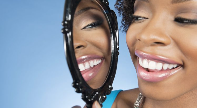 Close-up of a young woman looking at herself in mirror and smiling over colored background